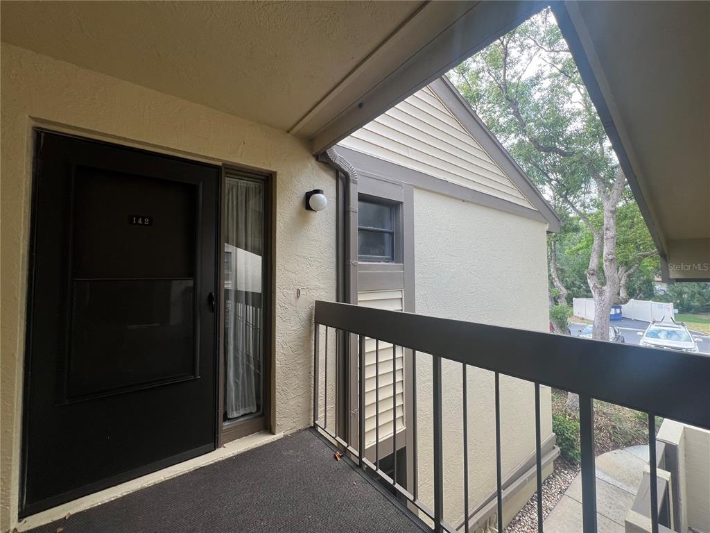 An outdoor balcony features a dark screen door, a metal railing, and beige stucco walls — 142 WOODLAKE WYNDE UNIT 48, OLDSMAR, FL