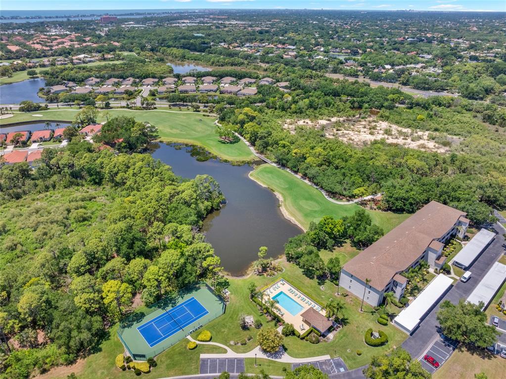 An aerial view shows a tennis court, swimming pool, pond, and apartment building near woods — 8750 OLDE HICKORY AVENUE UNIT 9308, SARASOTA, FL