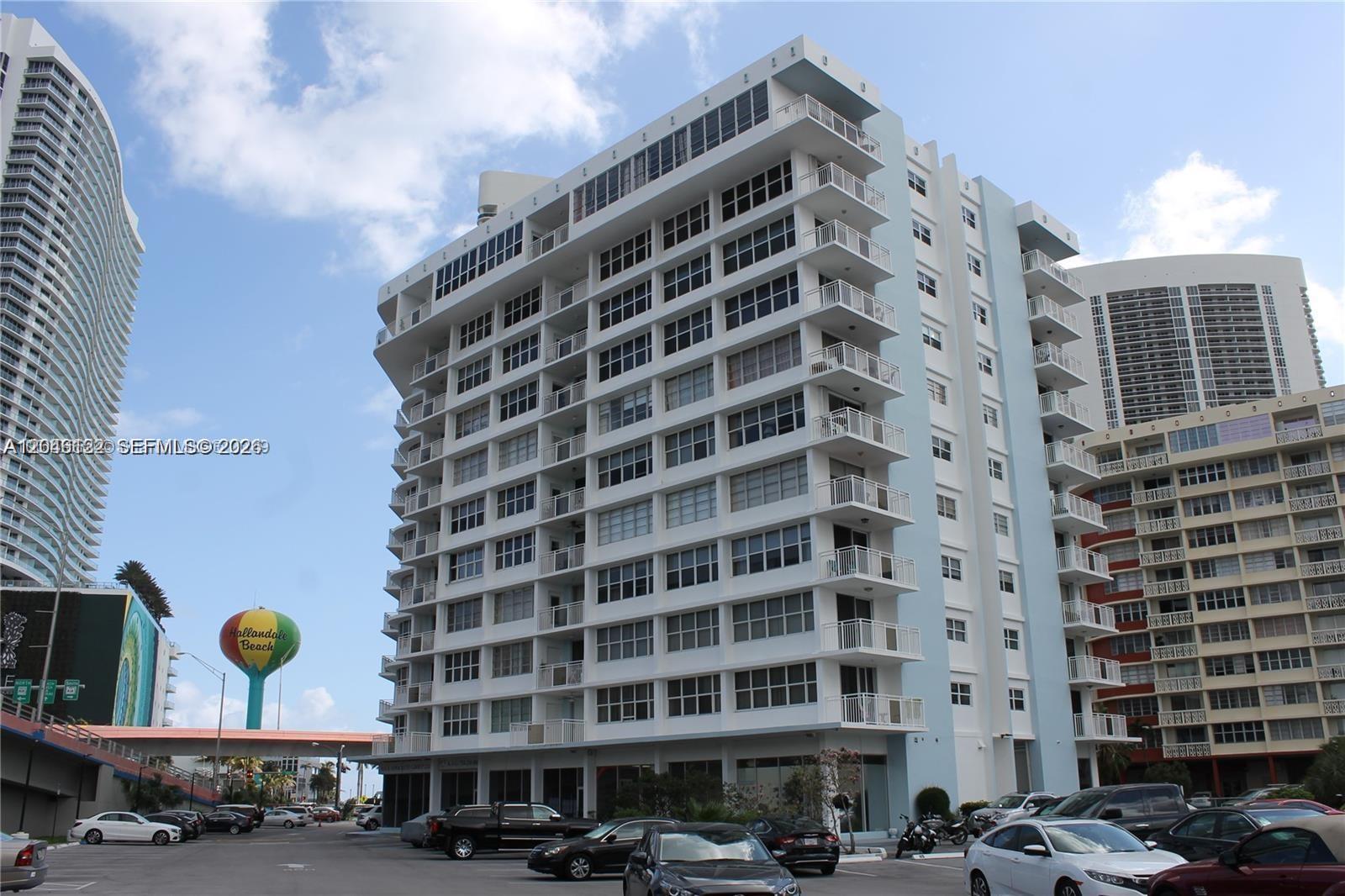 A multi-story residential building with balconies stands in a parking lot near a water tower — HALLANDALE BEACH, FL