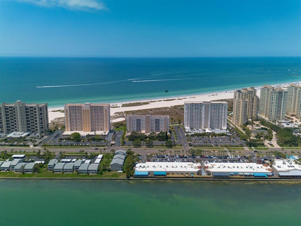 An aerial view shows high-rise beachfront condominiums along a sandy coastline with a nearby bay — 1250 GULF BOULEVARD UNIT 706, CLEARWATER BEACH, FL