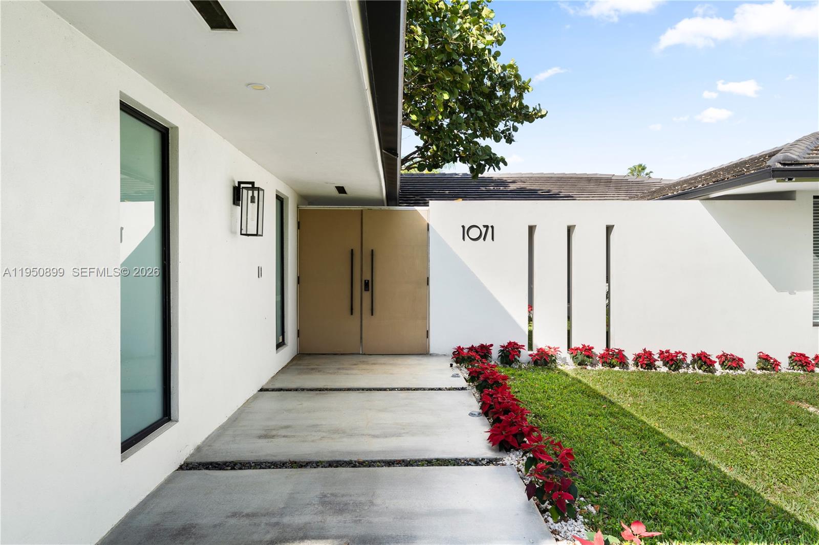A modern home entrance features a tan double door, concrete walkway, and red poinsettia landscaping — 1071 NE 83rd St, Miami FL 33138, MIAMI, FL