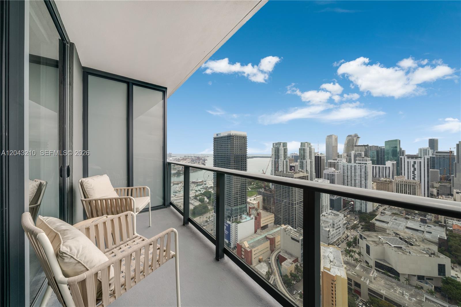 A high-rise balcony features two beige chairs overlooking a dense urban skyline and water — 159 NE 6th St # 4212, Miami FL 33132, MIAMI, FL