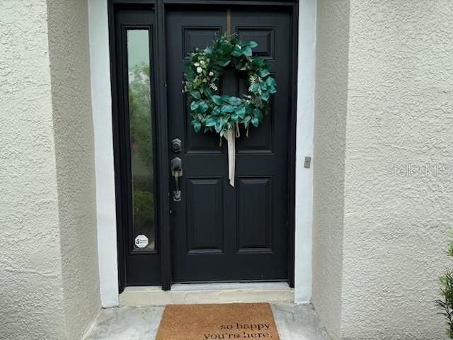 A black front door with a green wreath and a welcome mat sits in a stucco entryway — 8546 GREAT EGRET TRACE, NEW PORT RICHEY, FL