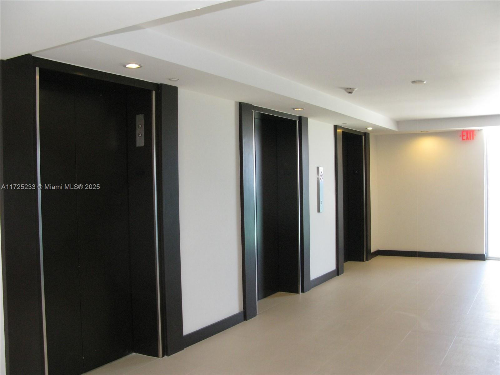A hallway features three black elevator doors set into white walls with recessed lighting — 5701 Collins Ave # 1011, Miami Beach FL 33140, MIAMI BEACH, FL