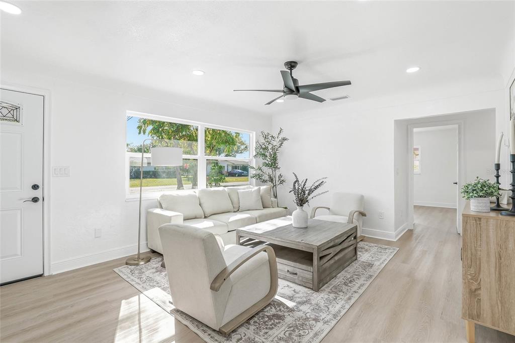 A bright living room features white walls, light wood-look flooring, a sofa, and two armchairs — 4720 MOHEGAN ROAD, ST PETERSBURG, FL