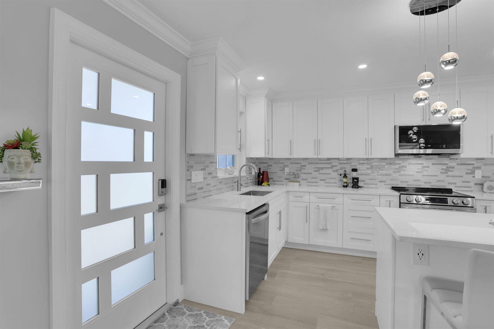A modern kitchen features white cabinetry, quartz countertops, a tiled backsplash, and a glass-paneled door — 540 High Point Drive # A, Delray Beach FL 33445, DELRAY BEACH, FL