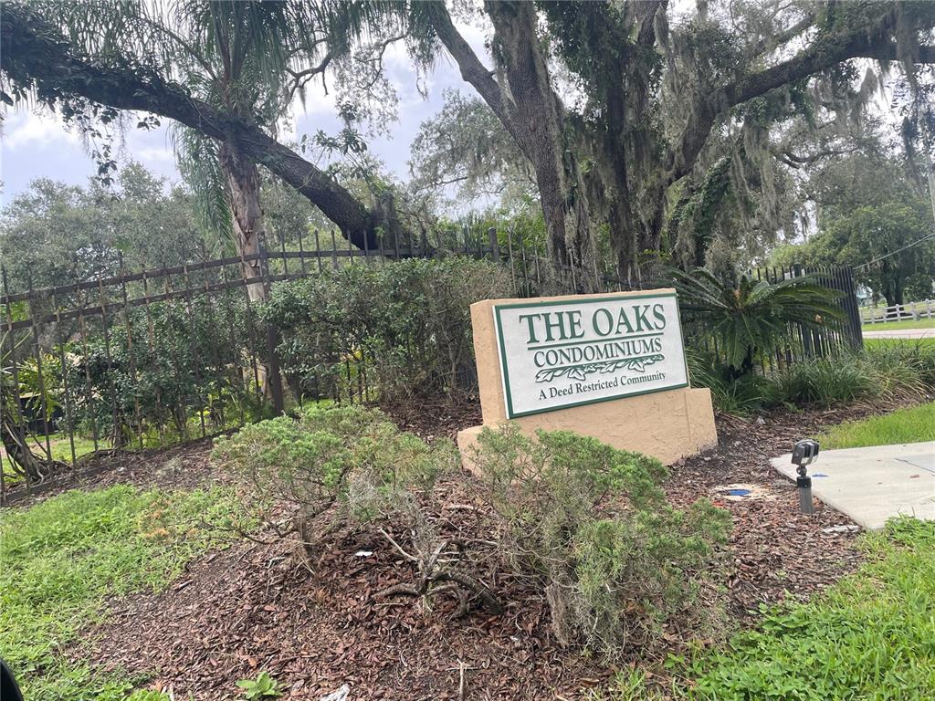 A sign for The Oaks Condominiums stands in front of a metal fence and trees — 14308 HANGING MOSS CIRCLE UNIT 101, TAMPA, FL