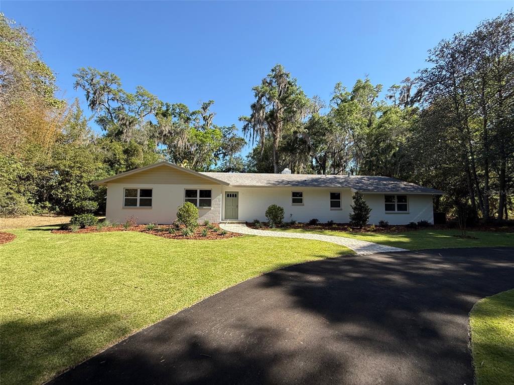 A single-story white house with a paved walkway and a curved driveway sits among trees — 4890 NW 20TH PLACE, GAINESVILLE, FL