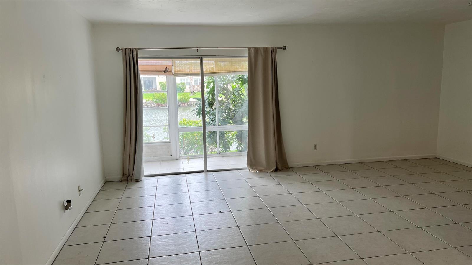 An empty room with light-colored tile flooring and a sliding glass door overlooking a canal — 1860 Robalo Drive # 104, Vero Beach FL 32960, VERO BEACH, FL