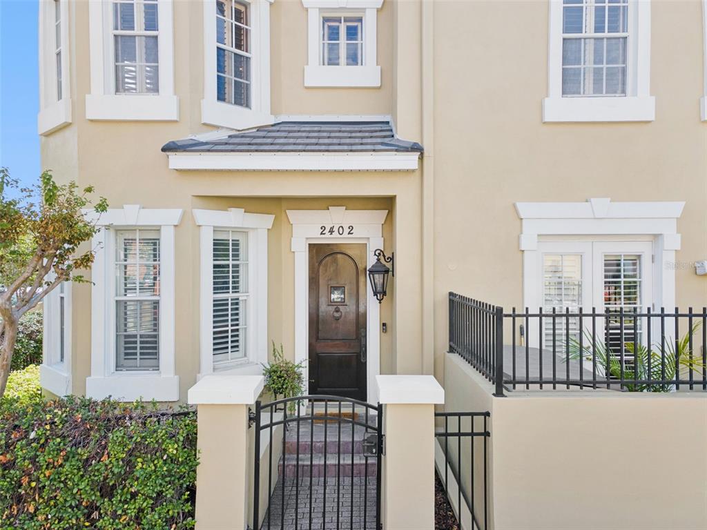 A beige townhouse exterior features a dark wood front door, black metal fencing, and windows — 2402 VICTORIA GARDENS LANE, TAMPA, FL