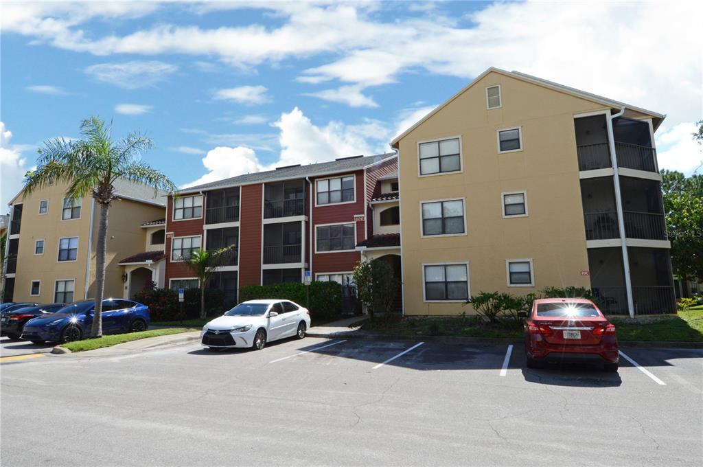 A three-story apartment building with tan and red siding stands behind a paved parking lot — 11901 4TH STREET N UNIT 6106, ST PETERSBURG, FL