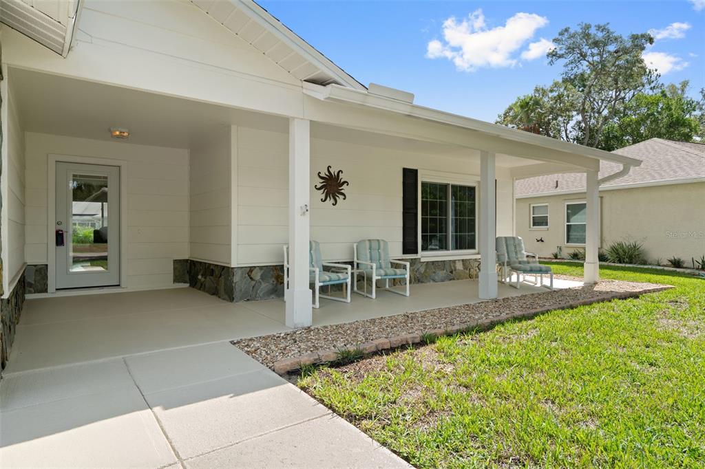 A covered front porch with white columns, stone accents, and three chairs sits beside a lawn — 2224 MERION COURT, SPRING HILL, FL