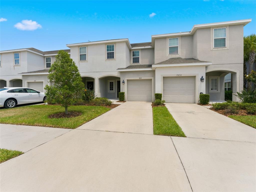 A multi-unit townhouse exterior features beige stucco walls, two garage doors, and a paved driveway — 7052 GRAND ELM DRIVE, RIVERVIEW, FL