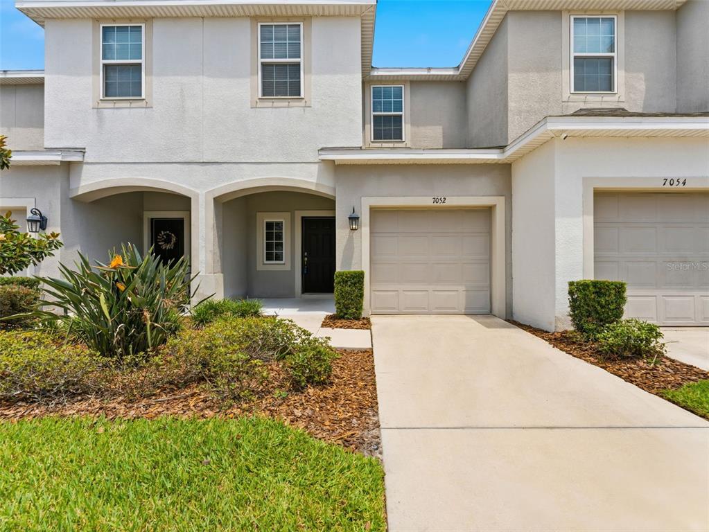A two-story townhouse exterior features a beige garage, a covered entryway, and landscaped front yard — 7052 GRAND ELM DRIVE, RIVERVIEW, FL