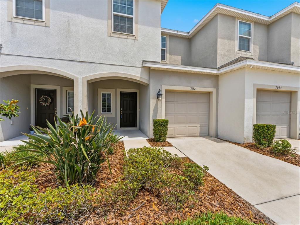 A two-story townhouse exterior features beige stucco, a single-car garage, and a landscaped front yard — 7052 GRAND ELM DRIVE, RIVERVIEW, FL