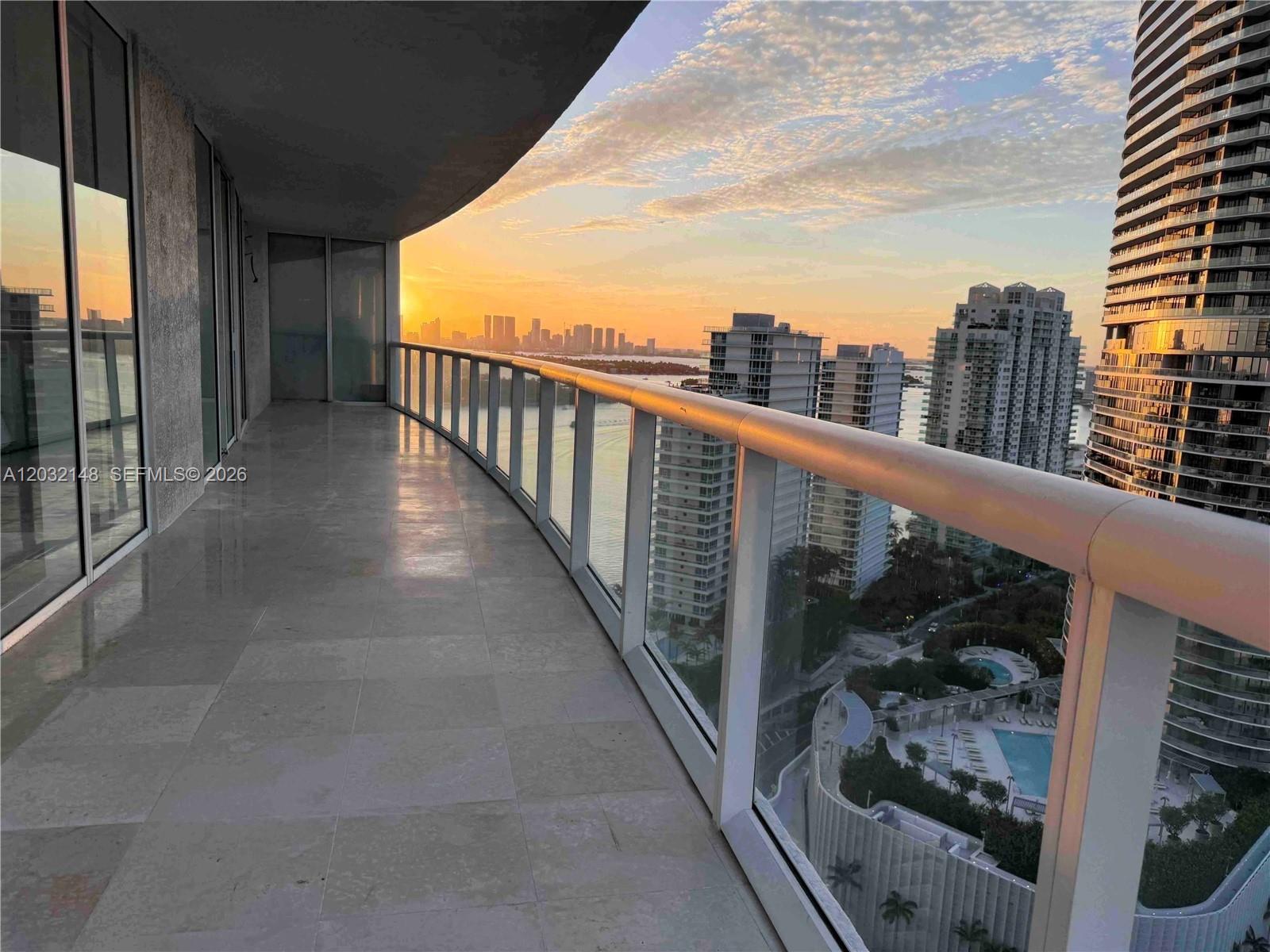 A curved balcony with glass railings overlooks a city skyline and water at sunset — 450 Alton Rd # 2610, Miami Beach FL 33139, MIAMI BEACH, FL