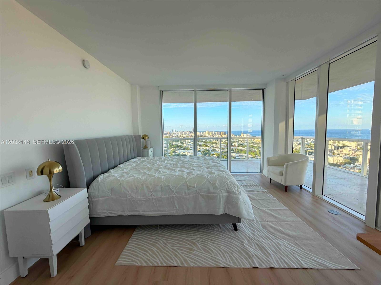 A modern bedroom features a gray upholstered bed, white nightstands, and floor-to-ceiling windows with ocean views — 450 Alton Rd # 2610, Miami Beach FL 33139, MIAMI BEACH, FL