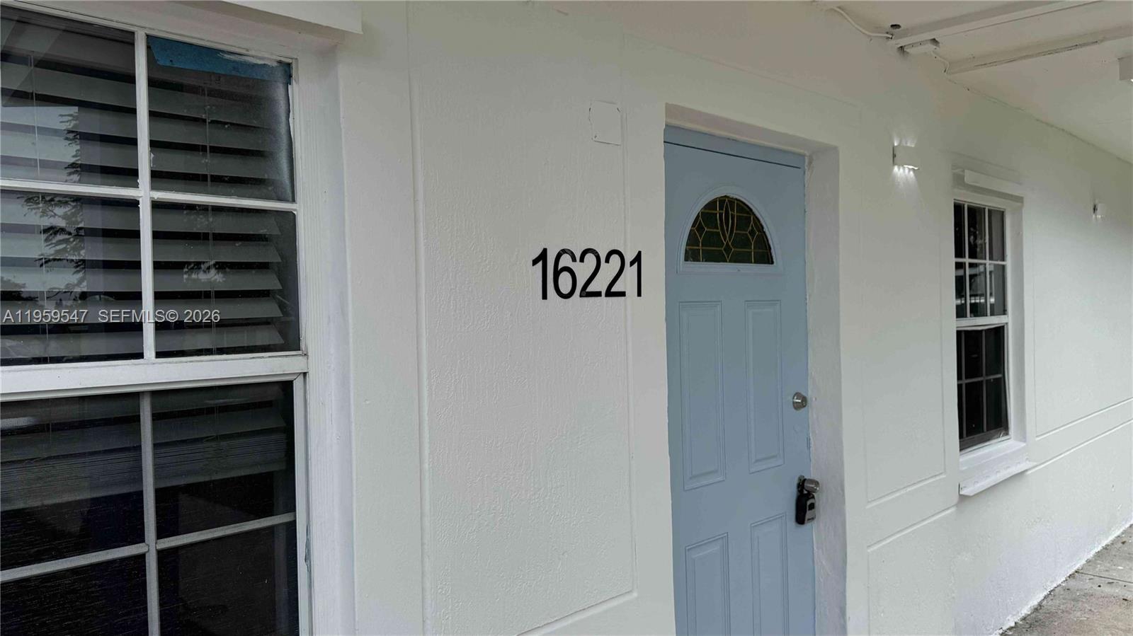 A white exterior wall features a light blue front door and a window with blinds — 16221 E Bunche Park Dr, Miami Gardens FL 33054, MIAMI GARDENS, FL
