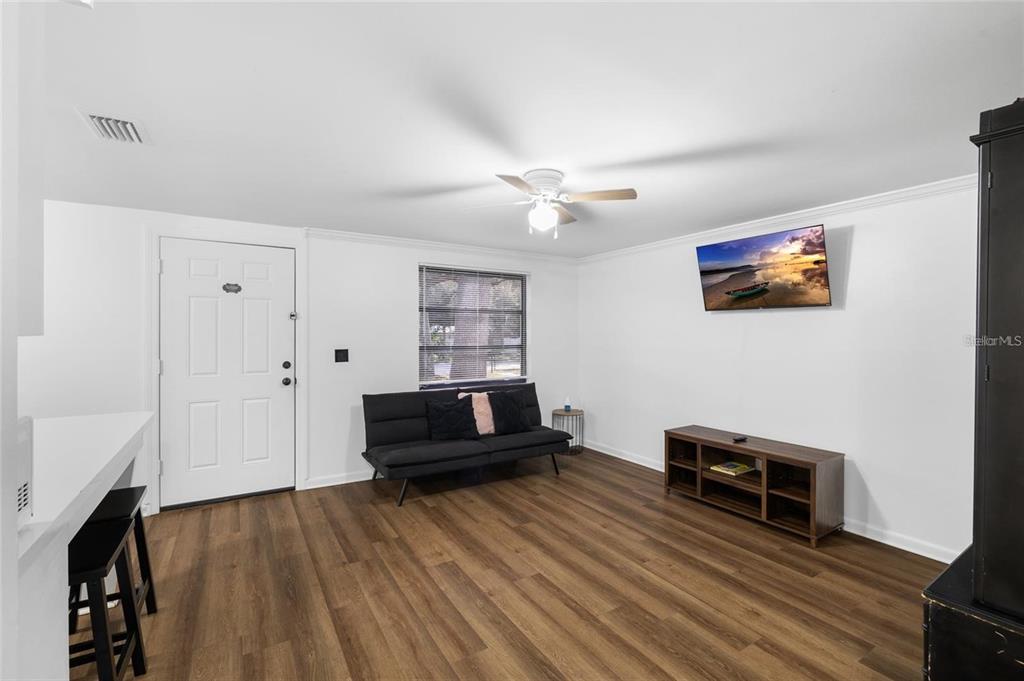 A living room features white walls, wood-look flooring, a black futon, and a wall-mounted television — 1220 E 142ND AVENUE UNIT B, TAMPA, FL