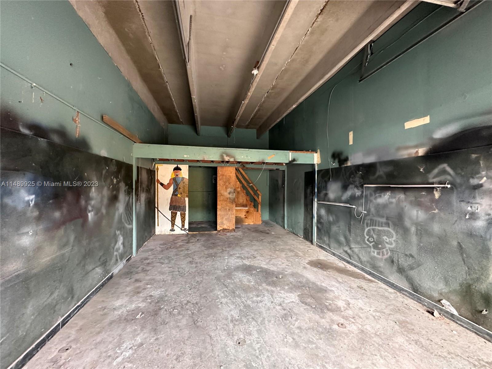 An unfinished, industrial-style room features concrete floors, dark walls with graffiti, and a wooden staircase — 5801-5807 Dawson St, Hollywood FL 33023, HOLLYWOOD, FL
