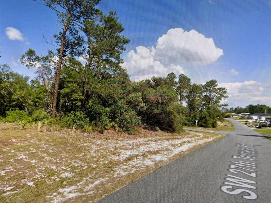 A vacant, wooded lot sits along a paved road under a blue sky — LOT 10 SW 27TH TERRACE ROAD, OCALA, FL