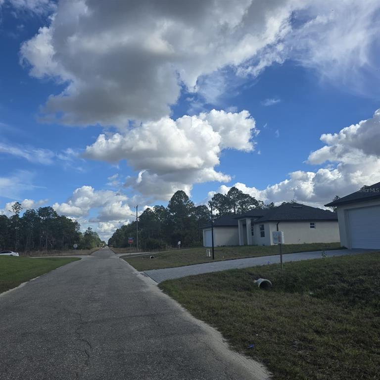 A paved street leads past a modern house toward a wooded area under a cloudy sky — 2603 E 13TH STREET, LEHIGH ACRES, FL