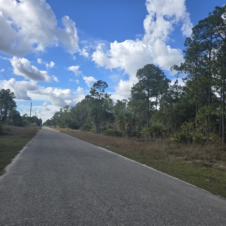 A paved road stretches past a wooded lot under a blue sky with clouds — 2603 E 13TH STREET, LEHIGH ACRES, FL