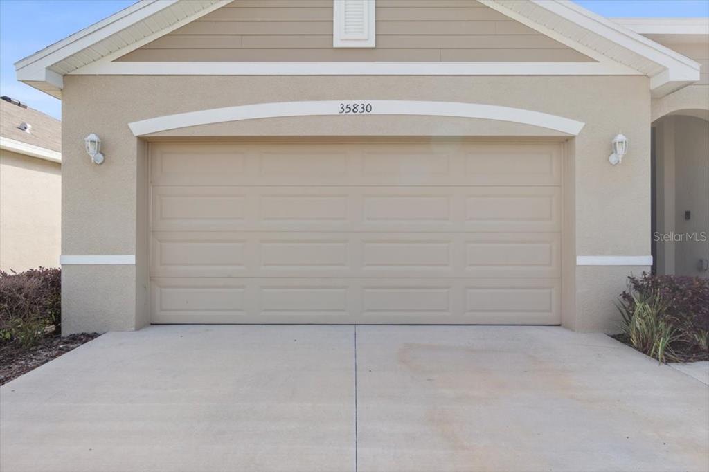 A beige house exterior features a two-car garage with a tan door and concrete driveway — 35830 ELLSWORTH PLACE, ZEPHYRHILLS, FL