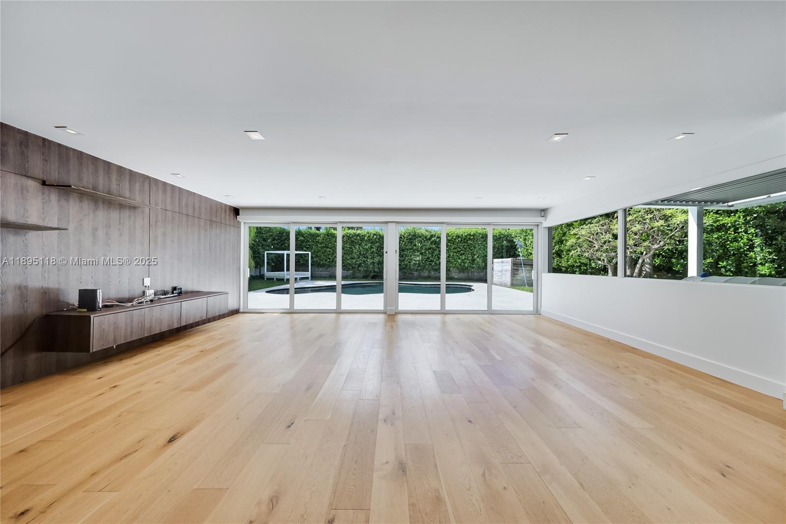 A spacious room features light wood flooring, a wood-paneled wall, and sliding glass doors — 5244 Alton Rd, Miami Beach FL 33140, MIAMI BEACH, FL