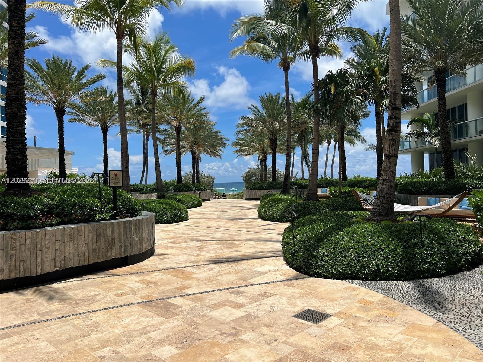 A paved walkway leads through a landscaped courtyard with palm trees toward the ocean — 6799 Collins Ave # 1501, Miami Beach FL 33141, MIAMI BEACH, FL