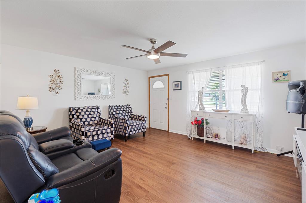 A living room features wood-look flooring, a ceiling fan, two patterned chairs, and a sofa — 5906 CYPRESS STREET, ZEPHYRHILLS, FL