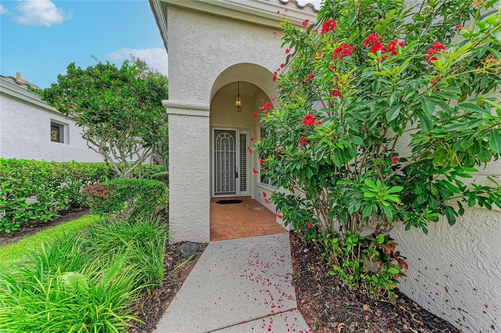 A concrete walkway leads to the arched entryway of a stucco home with red flowers — 3883 ALAMANDA DRIVE, SARASOTA, FL