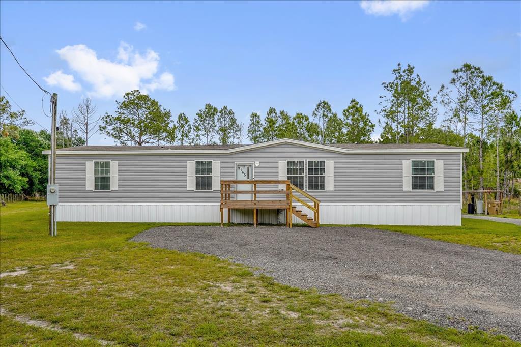 A single-story manufactured home with grey siding and a wooden front deck sits on gravel — 9165 LUNA DRIVE, ST CLOUD, FL