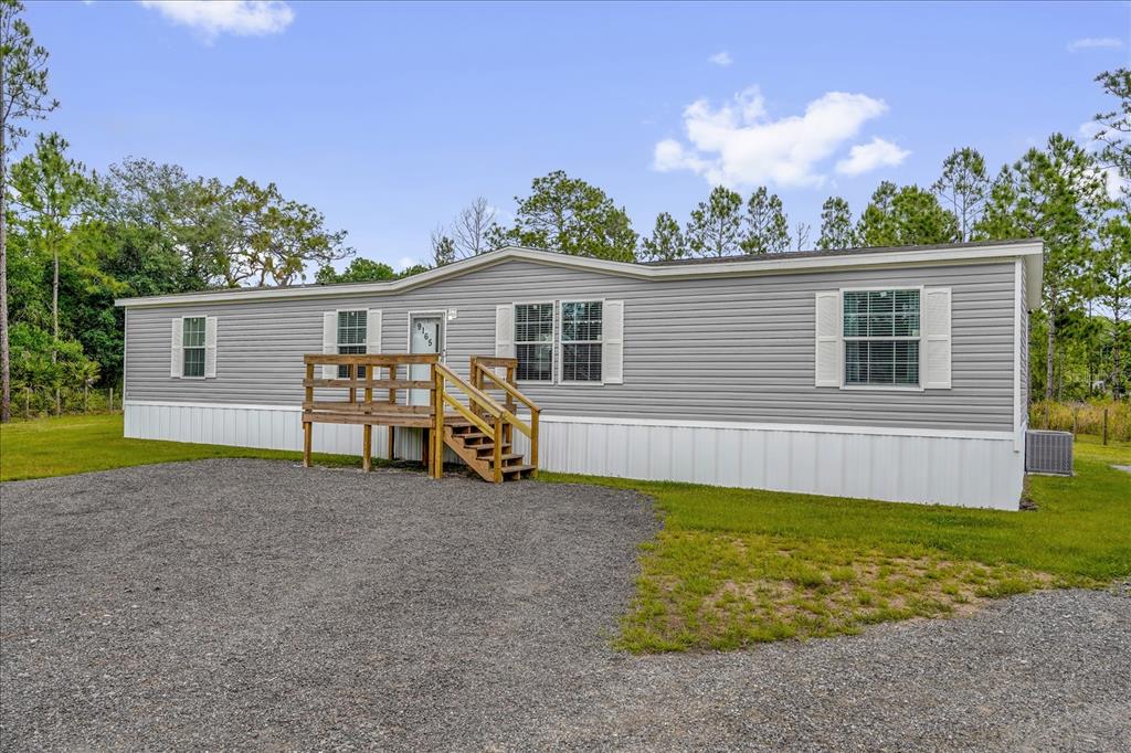 A single-story manufactured home with grey siding and a wooden deck sits on a gravel lot — 9165 LUNA DRIVE, ST CLOUD, FL