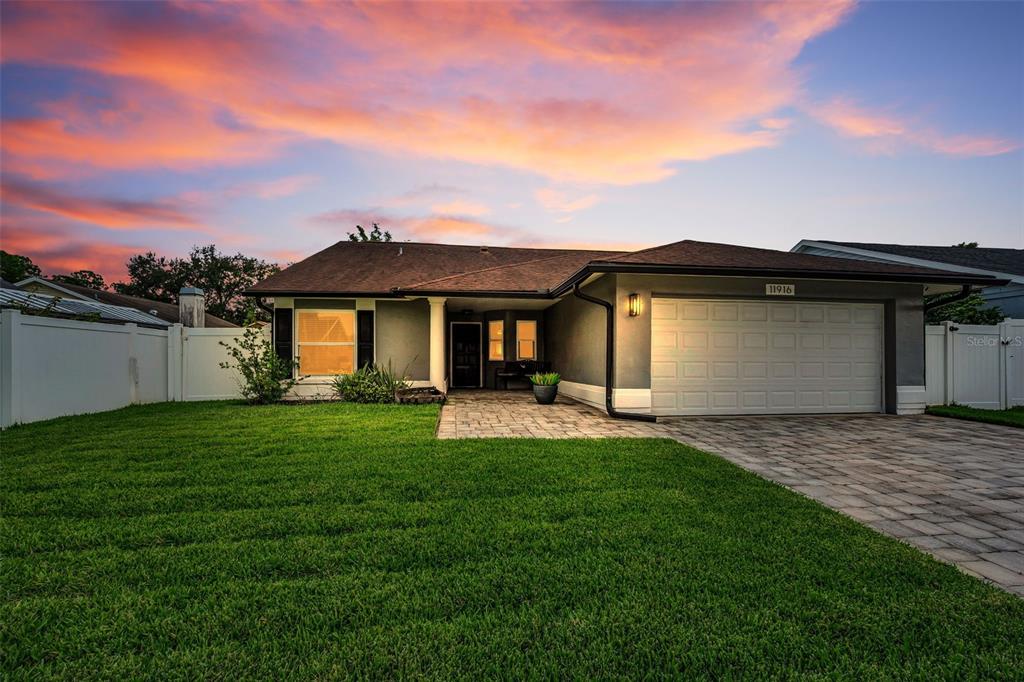 A single-story house exterior features a paved driveway, a green lawn, and a sunset sky — 11916 SNAPDRAGON ROAD, TAMPA, FL
