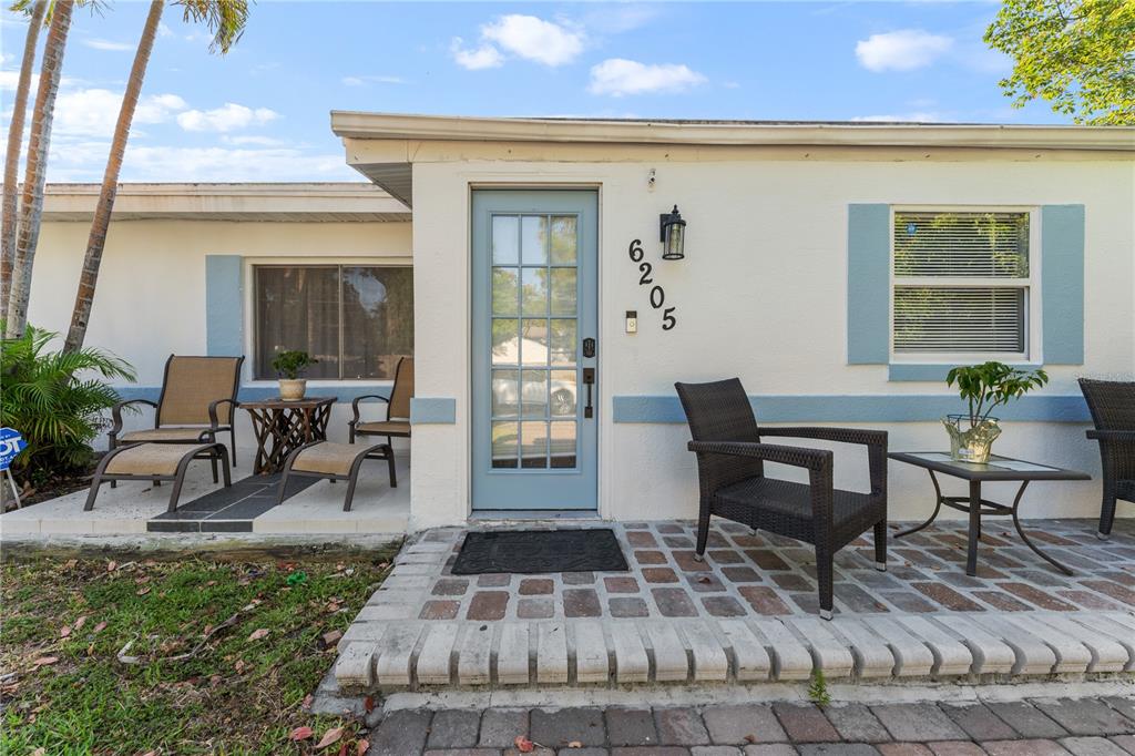 A light-colored house exterior features a blue front door, brick patio, and outdoor seating — 6205 65TH STREET N, PINELLAS PARK, FL