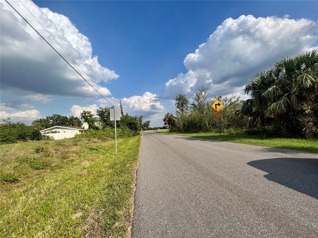 A paved road stretches forward under a blue sky with clouds, flanked by grassy areas — 27430 SAN MARCO DRIVE, PUNTA GORDA, FL
