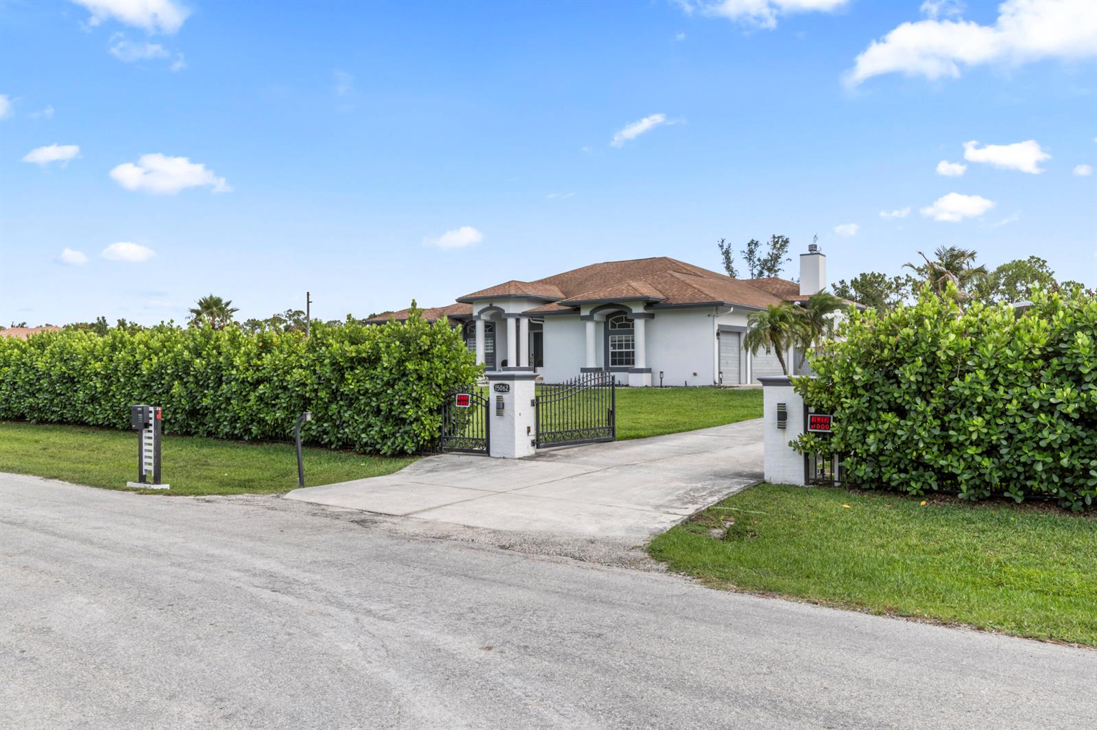 A single-story house with a gated driveway is surrounded by tall, dense green hedges — 15062 N 73rd St Street N, FL 33470, FL