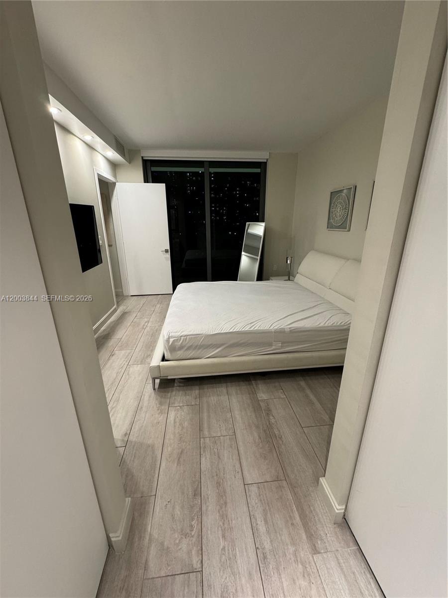 A bedroom features light wood-look tile flooring, a white upholstered bed, and a balcony door — 55 SW 9th St # 1204, Miami FL 33130, MIAMI, FL