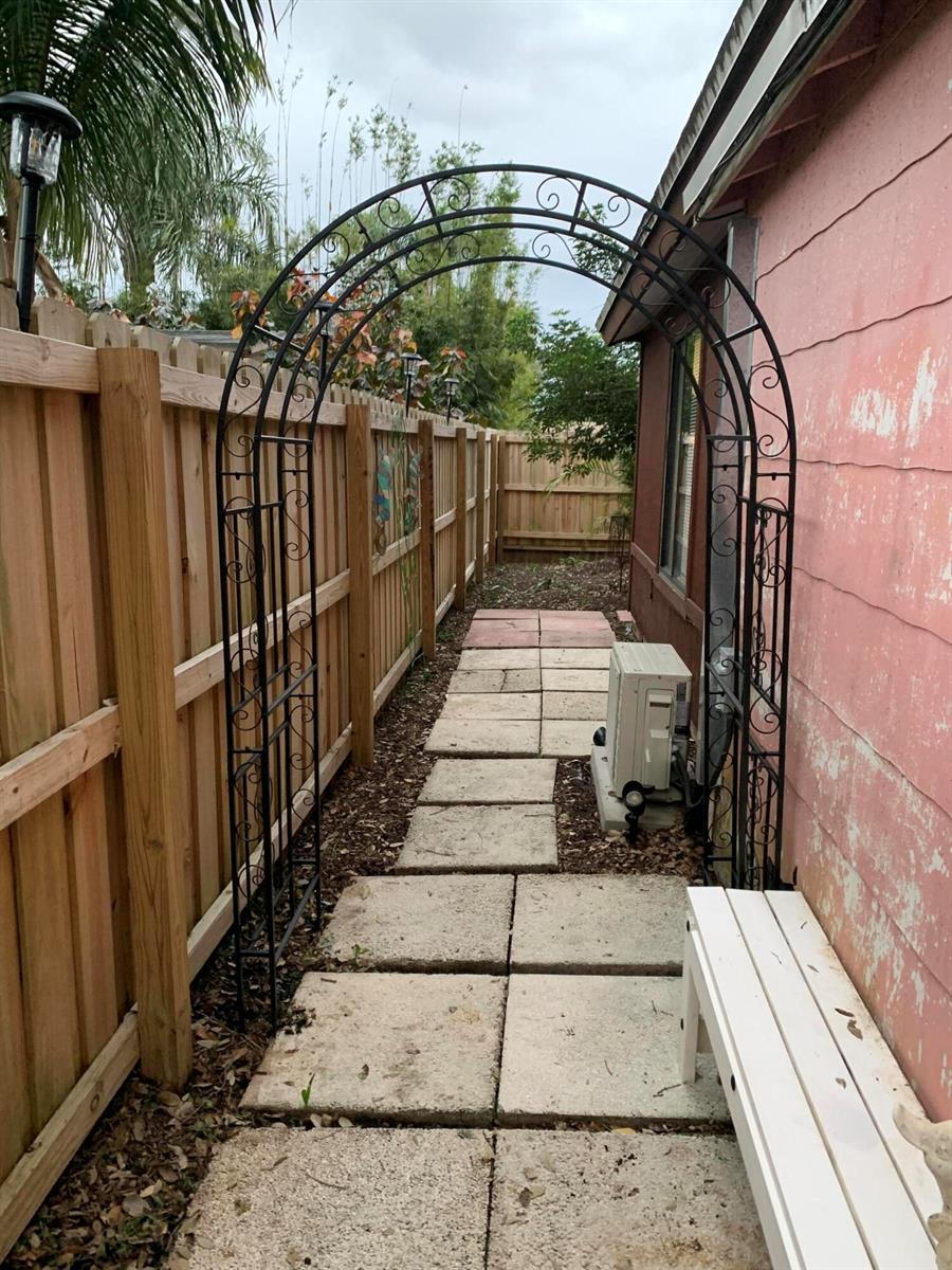 A metal garden arch stands over a stone walkway between a wooden fence and house — LAKE WORTH, FL