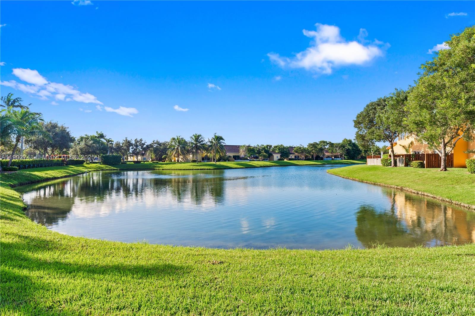 A calm lake is surrounded by green lawns, palm trees, and residential buildings under blue skies — 1398 NE 33rd Ave # 25, Homestead FL 33033, HOMESTEAD, FL