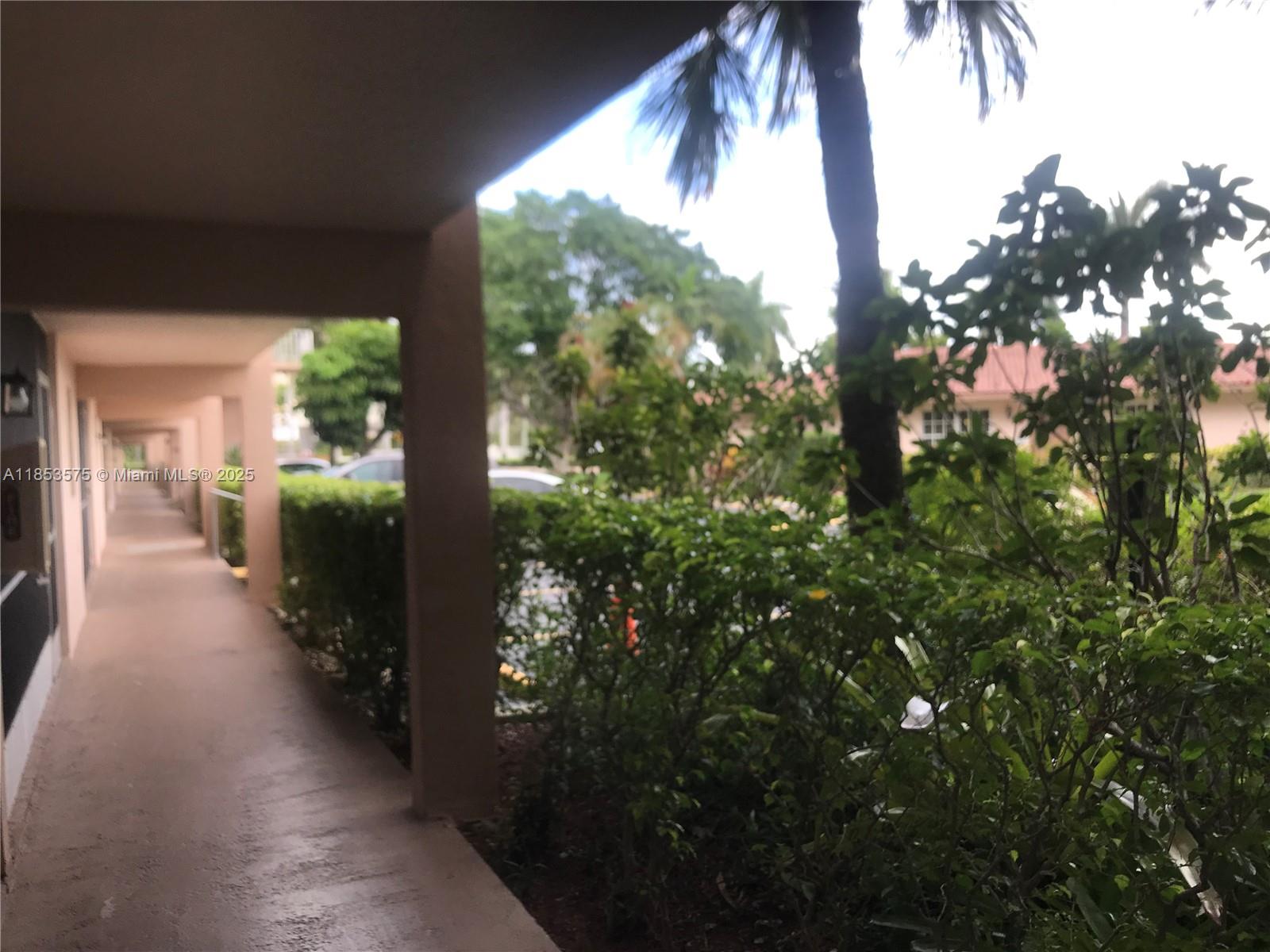 A covered outdoor walkway runs alongside a building with lush green landscaping and palm trees — 501 Blue Heron Dr # A, Hallandale Beach FL 33009, HALLANDALE BEACH, FL