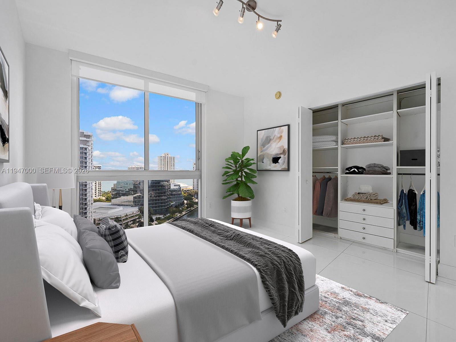 A modern bedroom features a white bed, a large window with city views, and an open closet — 495 Brickell Ave # 1801, Miami FL 33131, MIAMI, FL