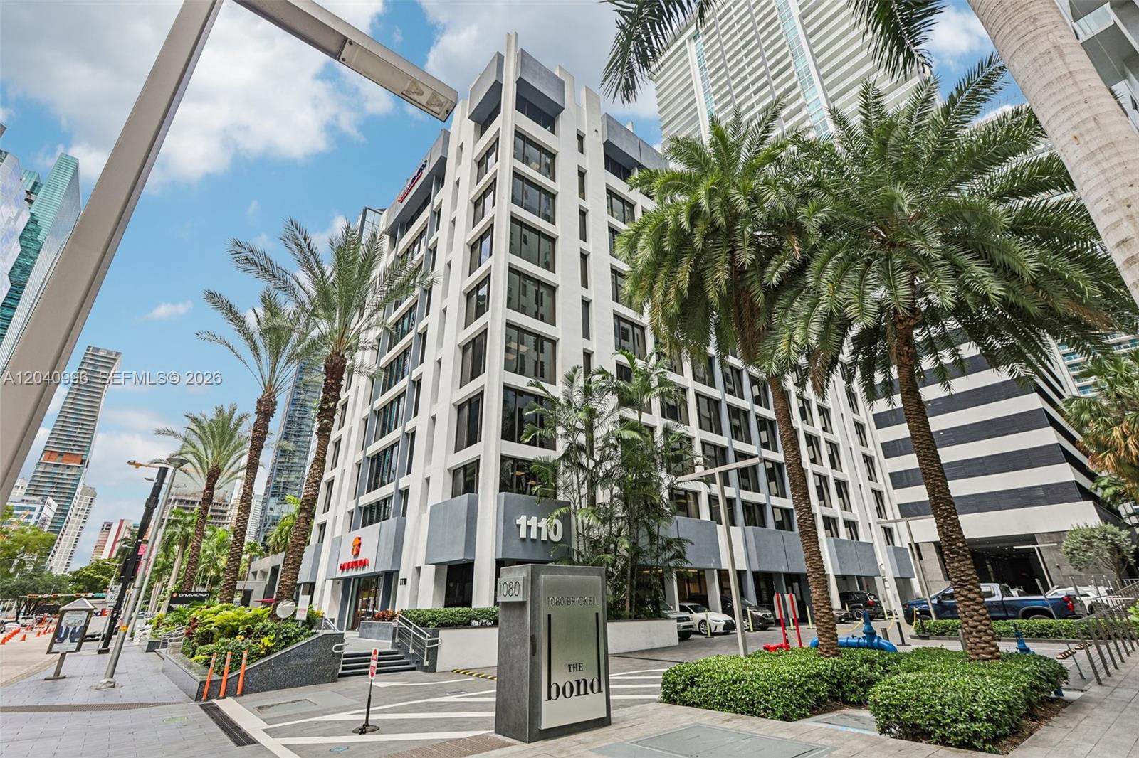 A low-angle exterior view of a white multi-story building with palm trees and signage — 1110 Brickell Ave # 512, Miami FL 33131, MIAMI, FL