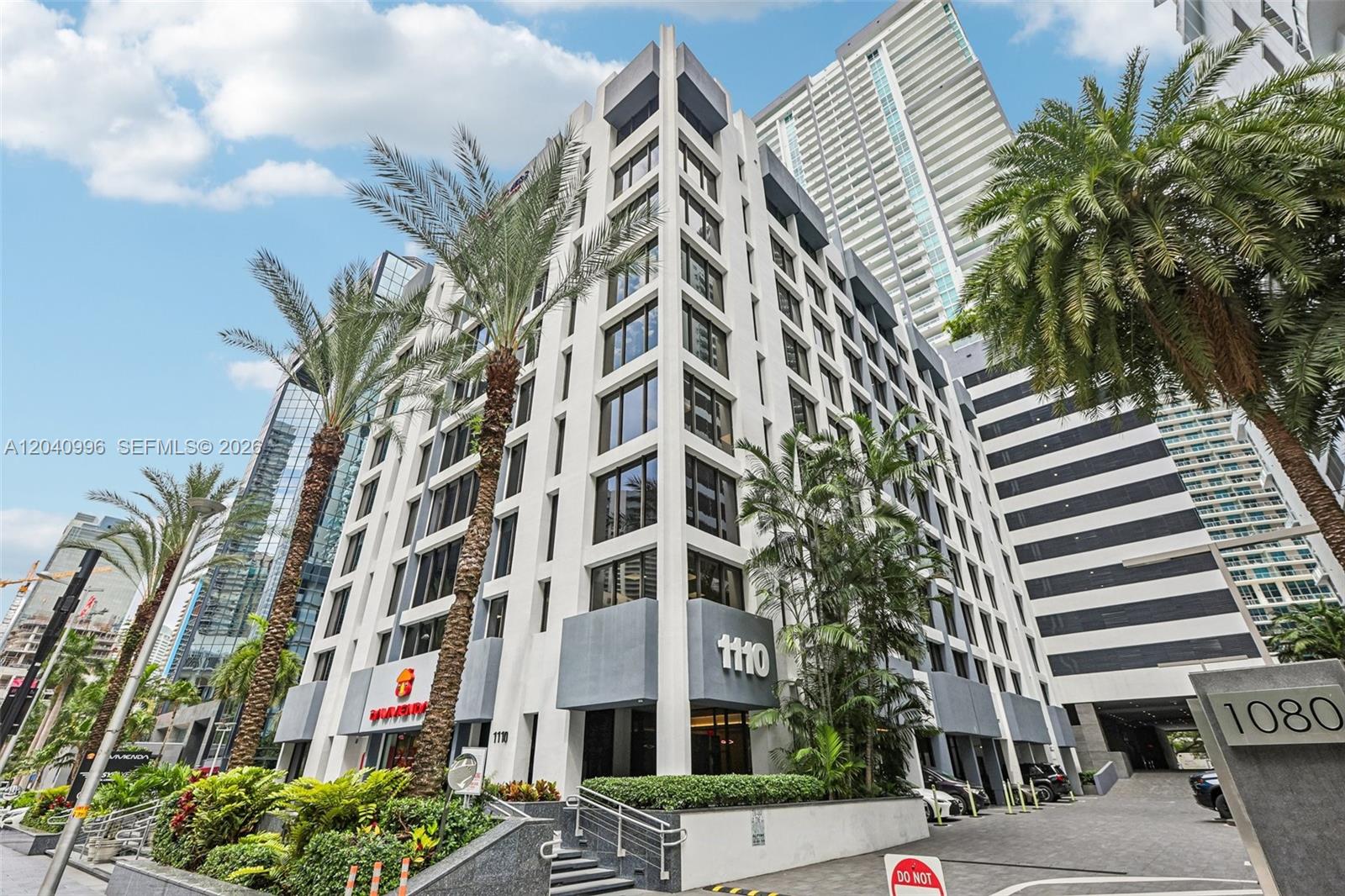 A low-angle exterior view of a multi-story white building with palm trees and signage — 1110 Brickell Ave # 512, Miami FL 33131, MIAMI, FL