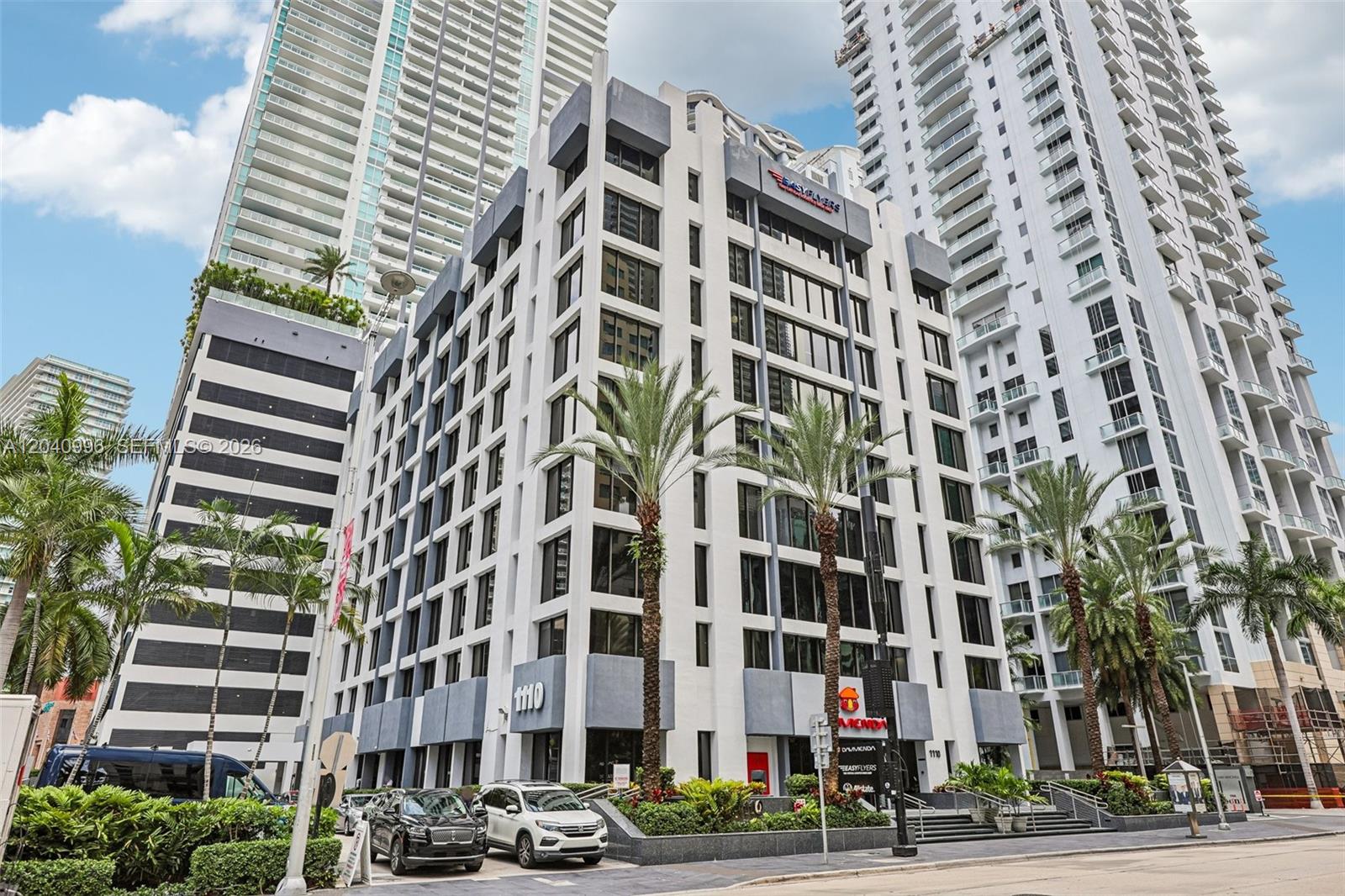A multi-story office building with a white facade and gray accents stands between two skyscrapers — 1110 Brickell Ave # 512, Miami FL 33131, MIAMI, FL