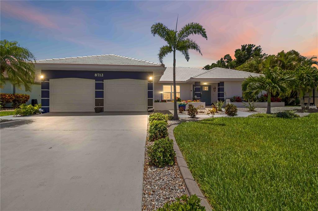 A single-story house with a two-car garage, white tile roof, and palm trees at dusk — 8712 53RD AVENUE W, BRADENTON, FL
