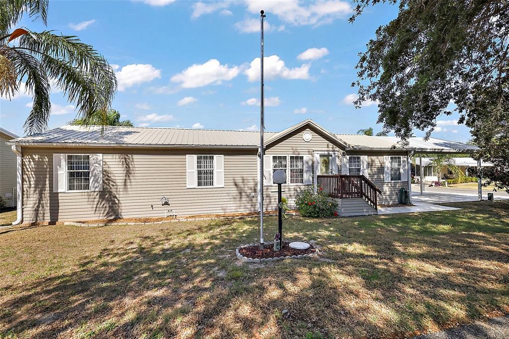 A single-story manufactured home with beige siding, a metal roof, and a front flagpole — 37945 MONTICELLO STREET, LEESBURG, FL