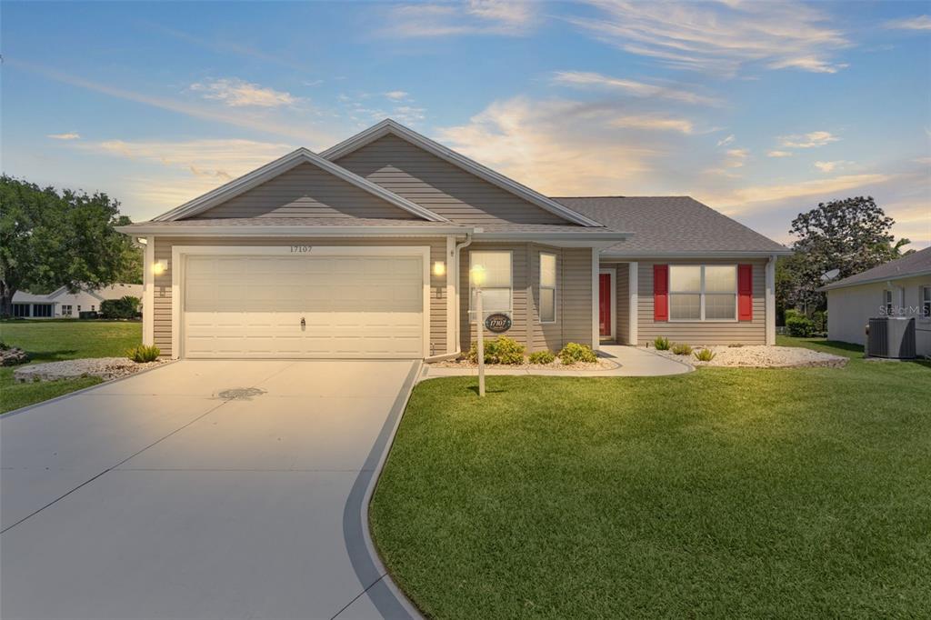 A single-story house with beige siding, a two-car garage, and red window shutters — 17107 SE 93RD EXETER COURT, THE VILLAGES, FL