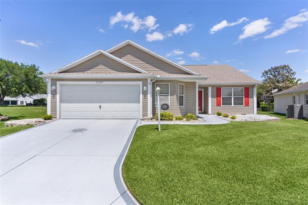 A single-story house with tan siding, a two-car garage, and a red front door — 17107 SE 93RD EXETER COURT, THE VILLAGES, FL